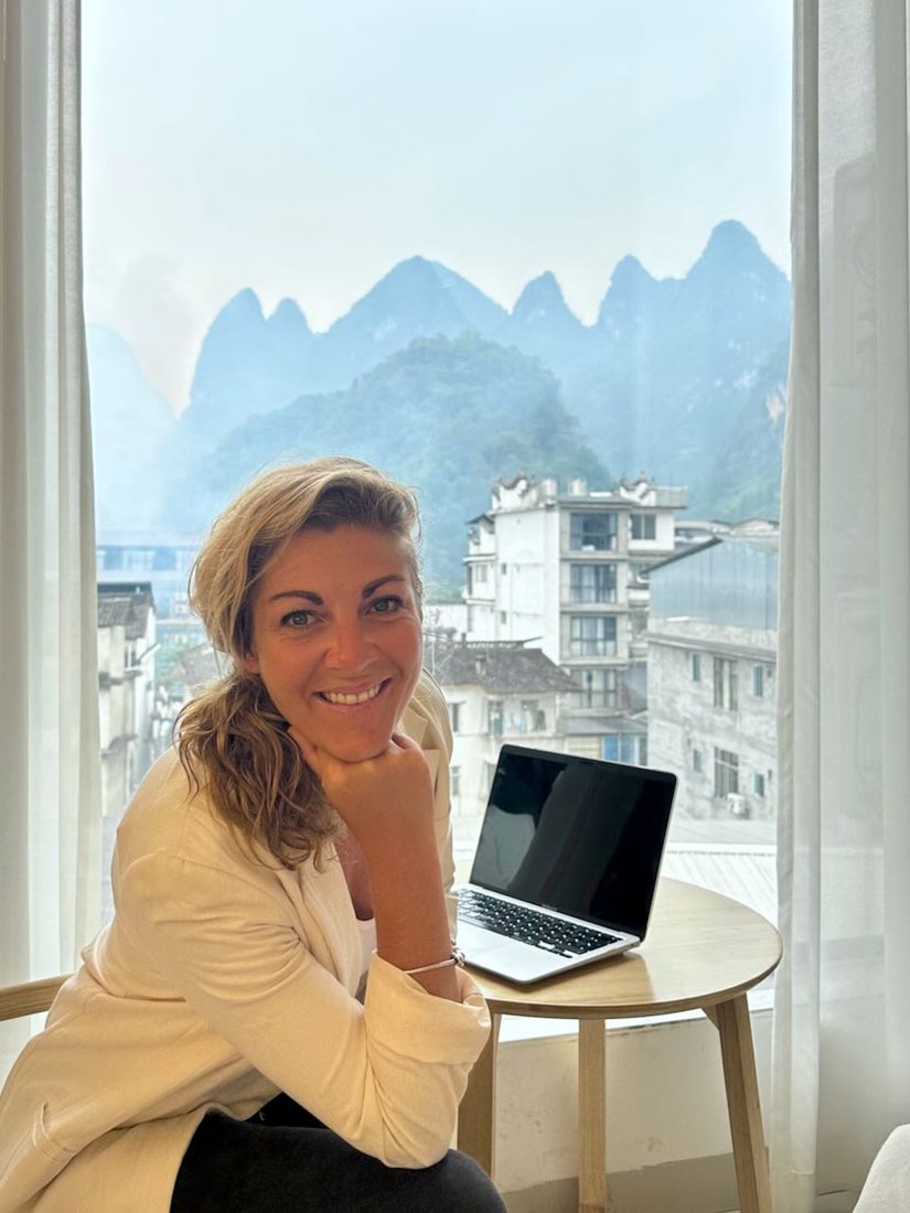 Verena em uma mesinha com o notebook, montanhas pela janela atrás dela