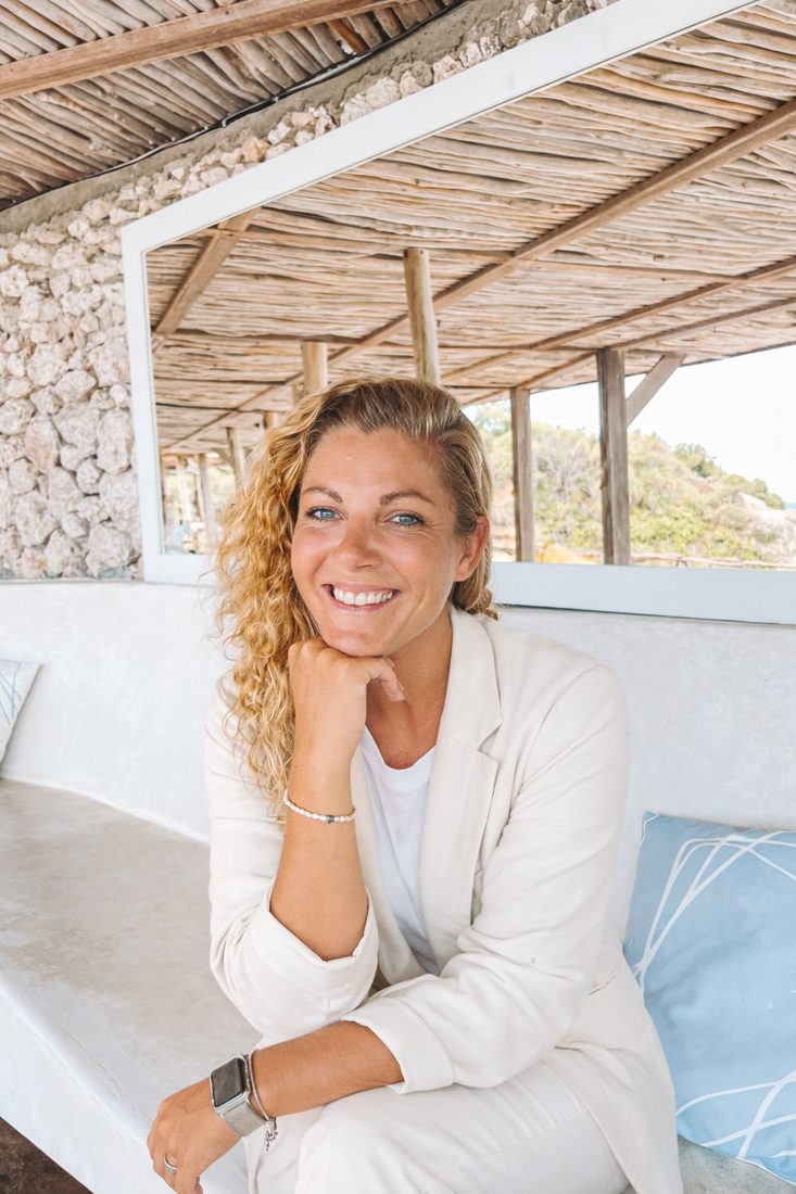 Verena sorrindo, sentada numa cabana de pedra na praia
